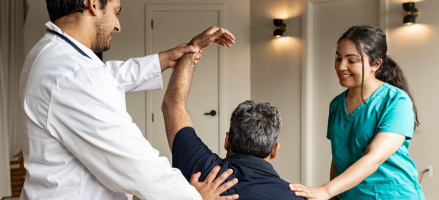 A man raises his arm as a Physiotherapist maneuvers his shoulder and a support worker reassures him
